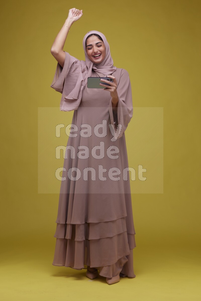 A woman Playing Games a Yellow Background wearing Brown Abaya with Hijab