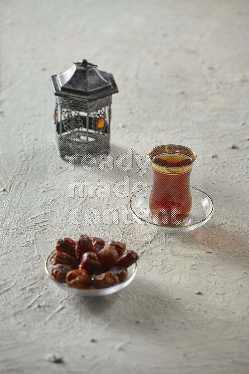 A silver lantern with different drinks, dates, nuts, prayer beads and quran on textured white background