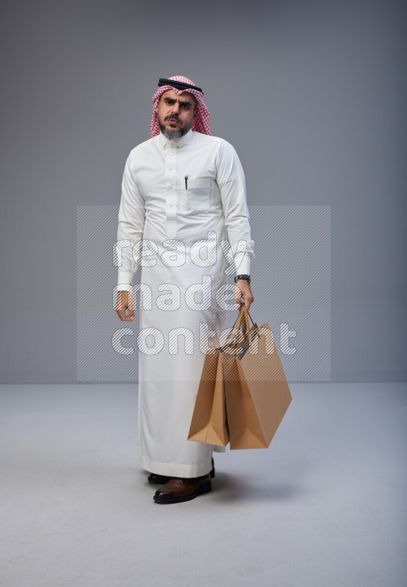 Saudi man Wearing Thob and red Shomag standing holding shopping bag on Gray background