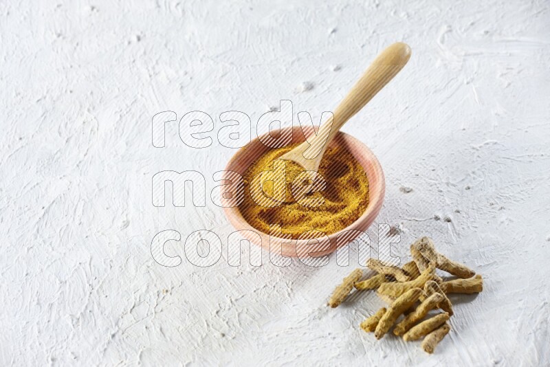 A wooden bowl and wooden spoon full of turmeric powder with dried turmeric fingers beside it on textured white flooring