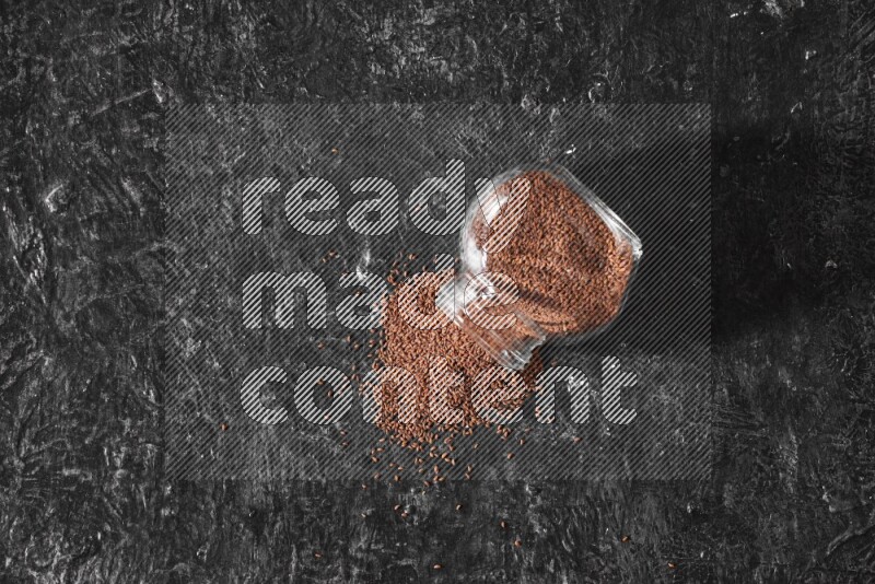 وعاء زجاجي مقلوب ممتلئ ببذور حب الرشاد وتناثرت البذور منه على أرضية سوداء