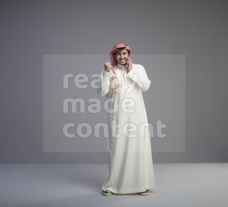 A Saudi man standing wearing thob and red shomag talking on phone on gray background