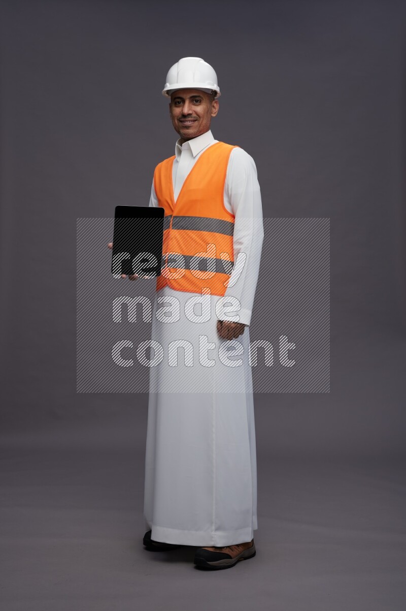 Saudi man wearing thob with engineer vest standing showing tablet to camera on gray background