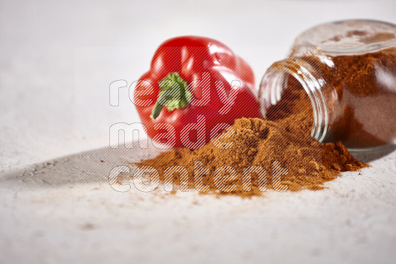 A glass jar full of ground paprika powder flipped with some spilling powder on white background