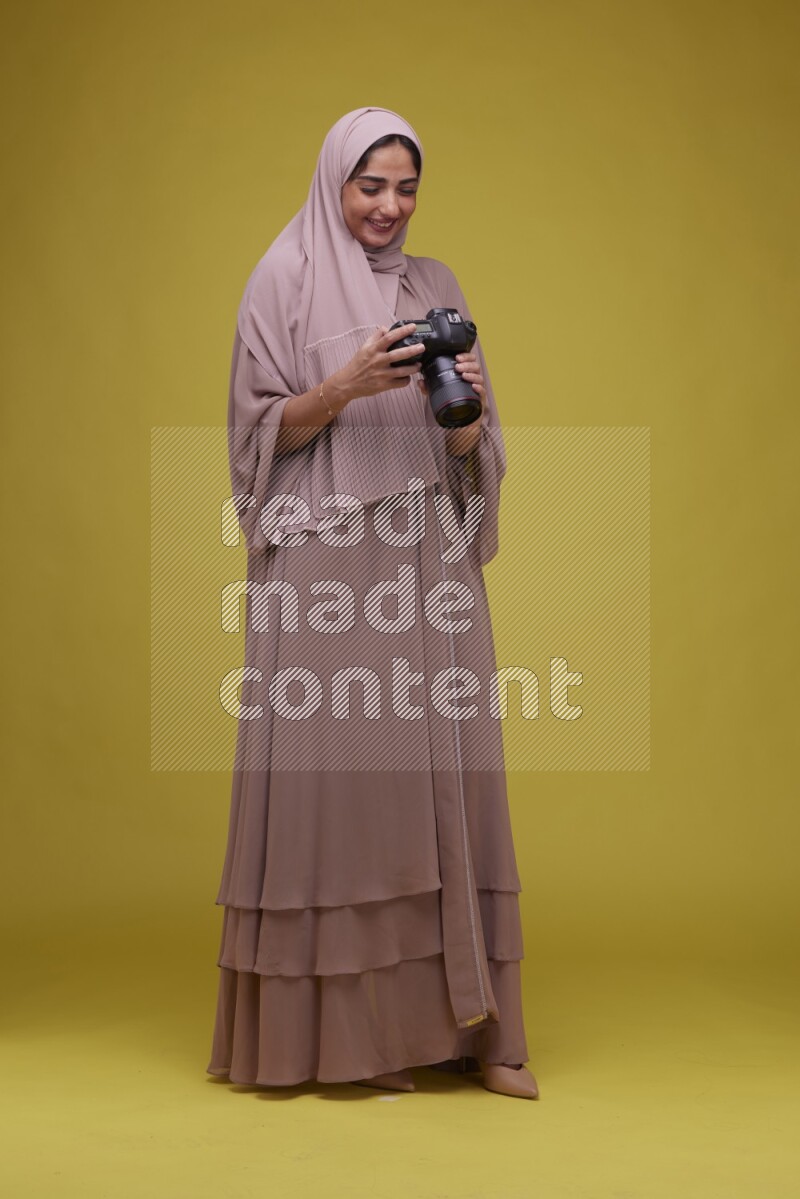 A woman Holding a Camera on a Yellow Background wearing Brown Abaya with Hijab