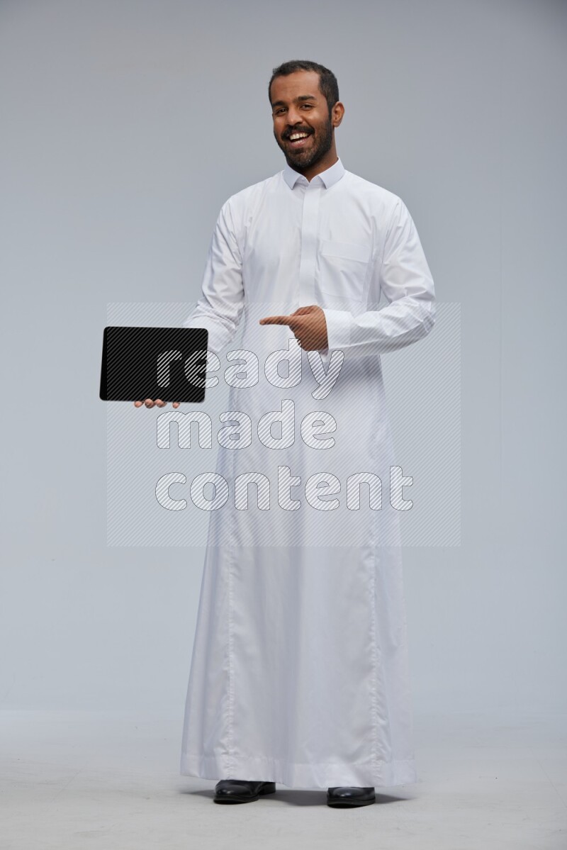 Saudi man wearing Thob standing showing tablet to camera on Gray background