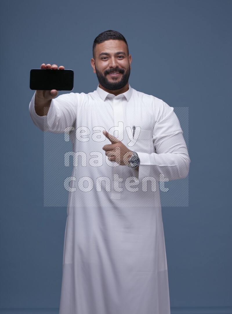 A man Showing his phone in Blue Background wearing Saudi Thob
