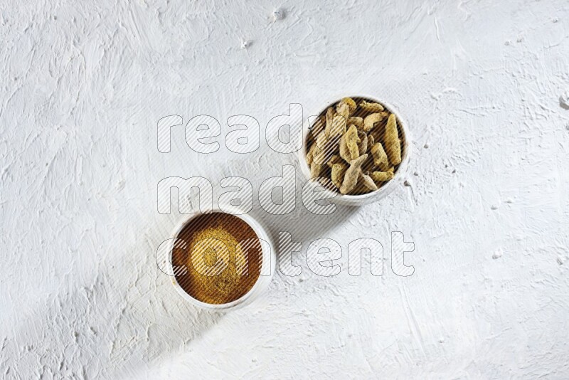 2 beige pottery bowl full of turmeric powder and dried turmeric whole fingers on textured white flooring