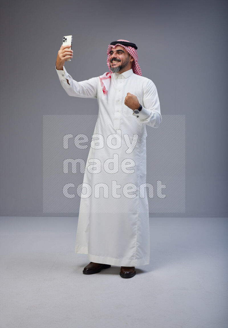 Saudi man Wearing Thob and red Shomag standing taking selfie on Gray background