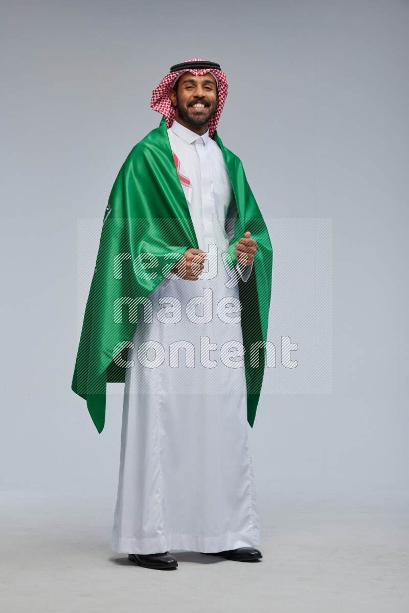 Saudi man Wearing Thob and shomag standing holding Saudi flag on Gray background