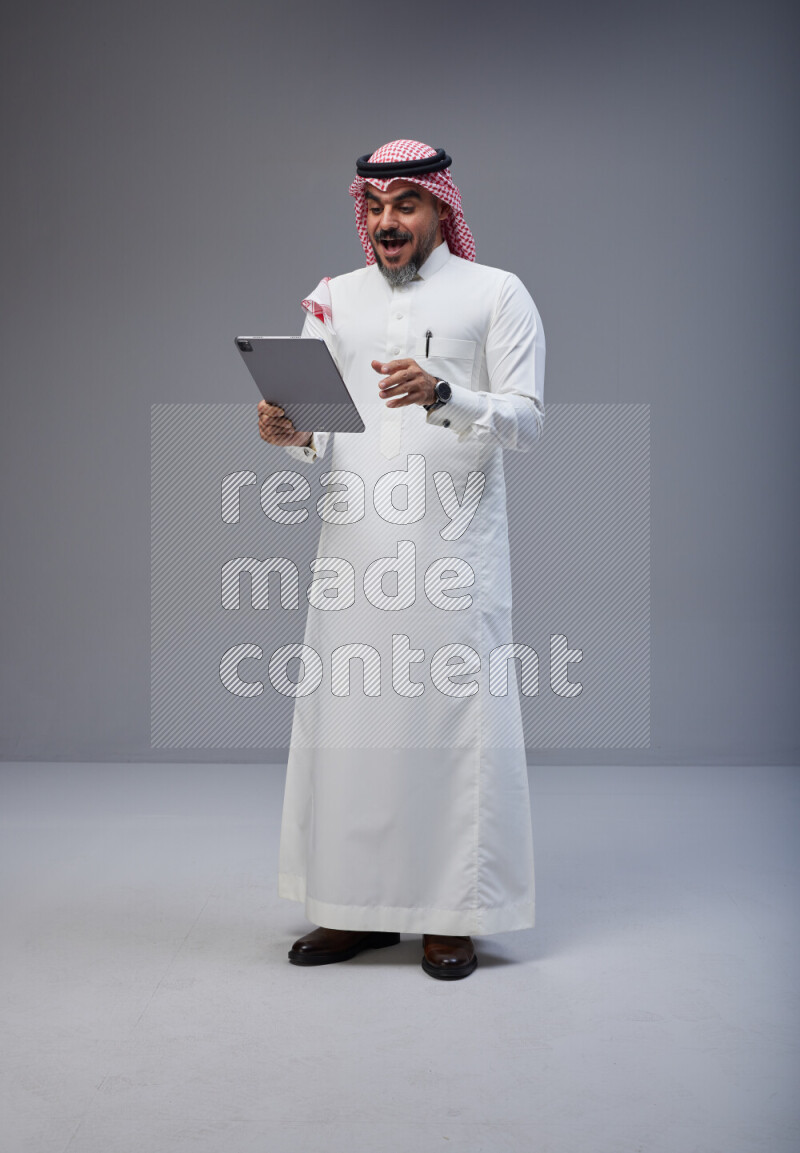 Saudi man Wearing Thob and red Shomag standing working on tablet on Gray background