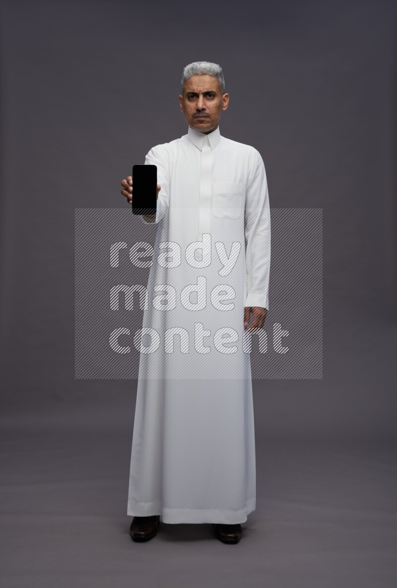 Saudi man wearing thob standing showing phone to camera on gray background