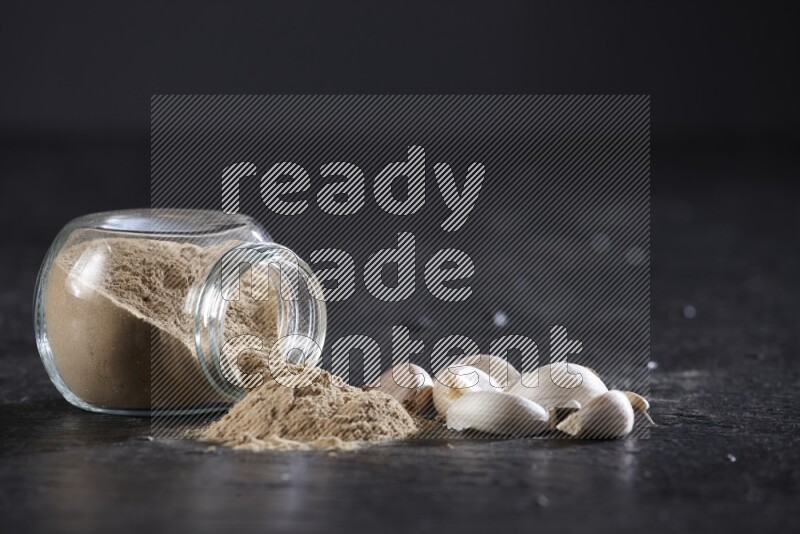 وعاء توابل زجاجي ممتلئ ببودرة الثوم مقلوب والبودرة متناثرة منه علي خلفية سوداء