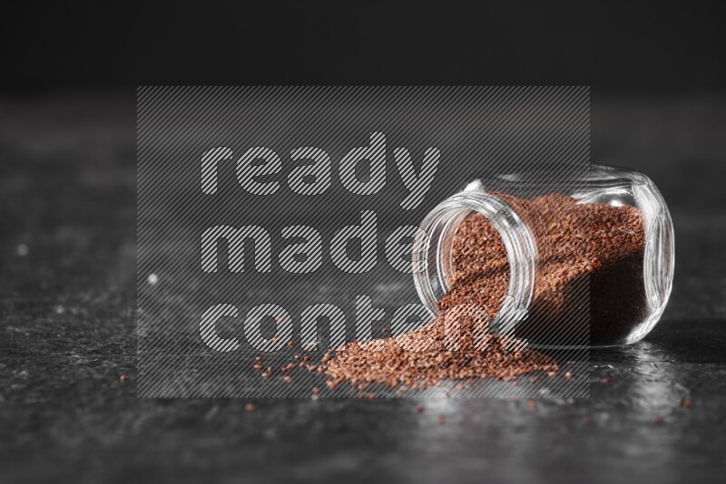 وعاء زجاجي مقلوب ممتلئ ببذور حب الرشاد وتناثرت البذور منه على أرضية سوداء