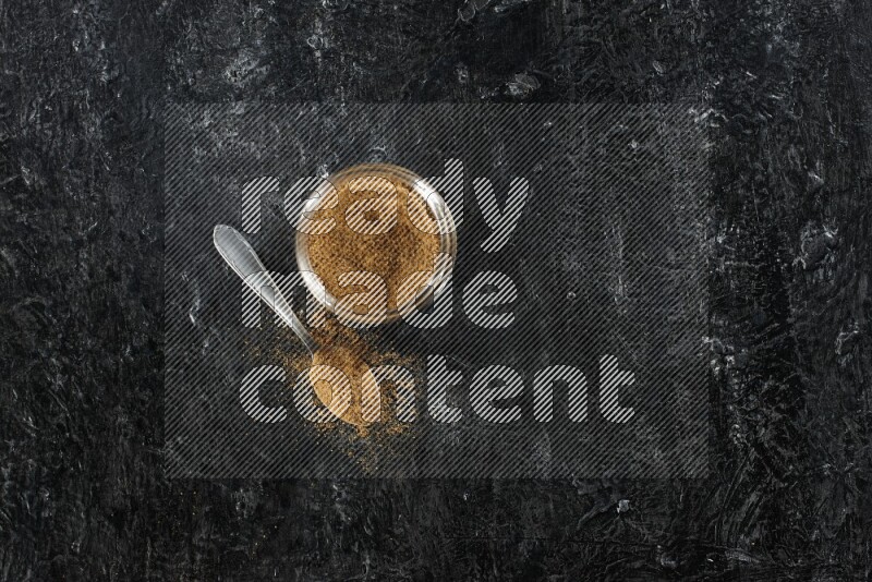 وعاء زجاجي للتوابل وملعقة معدنية ممتلئين ببودرة الكمون على خلفية سوداء