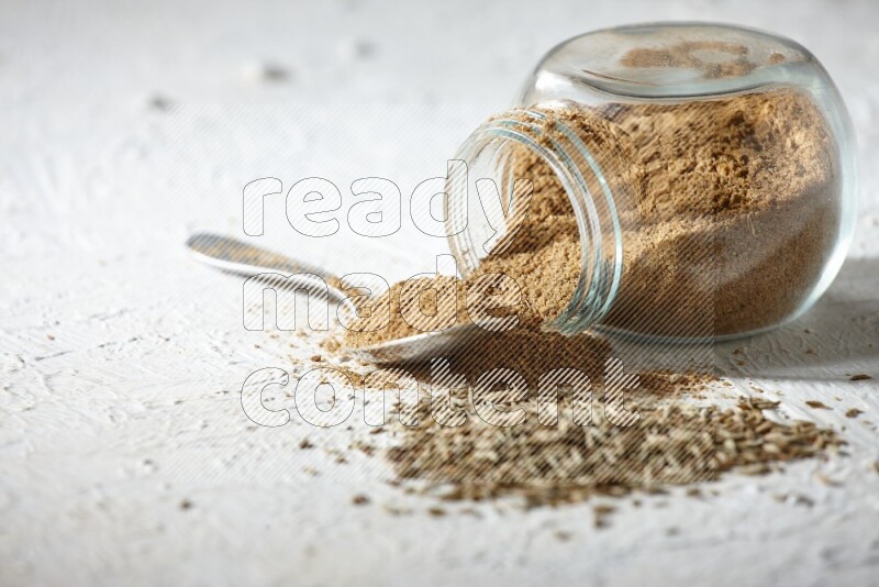 A glass spice jar and metal spoon full of cumin powder and the jar flipped and powder spilled out with cumin seeds on textured white flooring