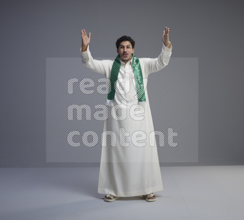 A Saudi man standing wearing thob and Saudi flag scarf on gray background