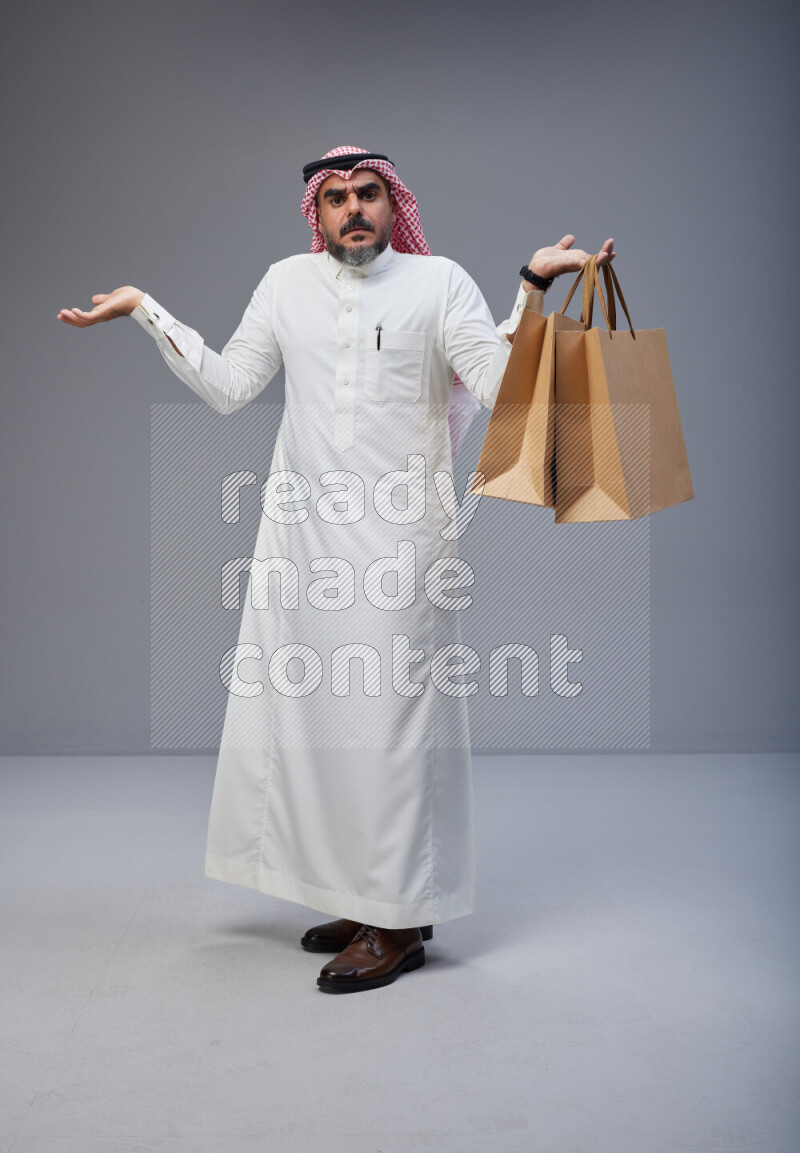 Saudi man Wearing Thob and red Shomag standing holding shopping bag on Gray background