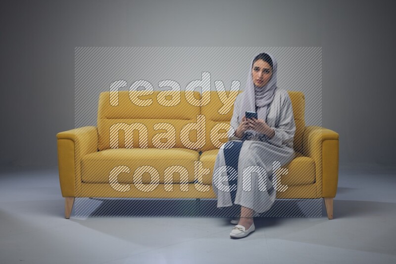 A Saudi woman wearing a light gray Abaya and white head scarf sitting on a yellow sofa and using her phone eye level on a grey background