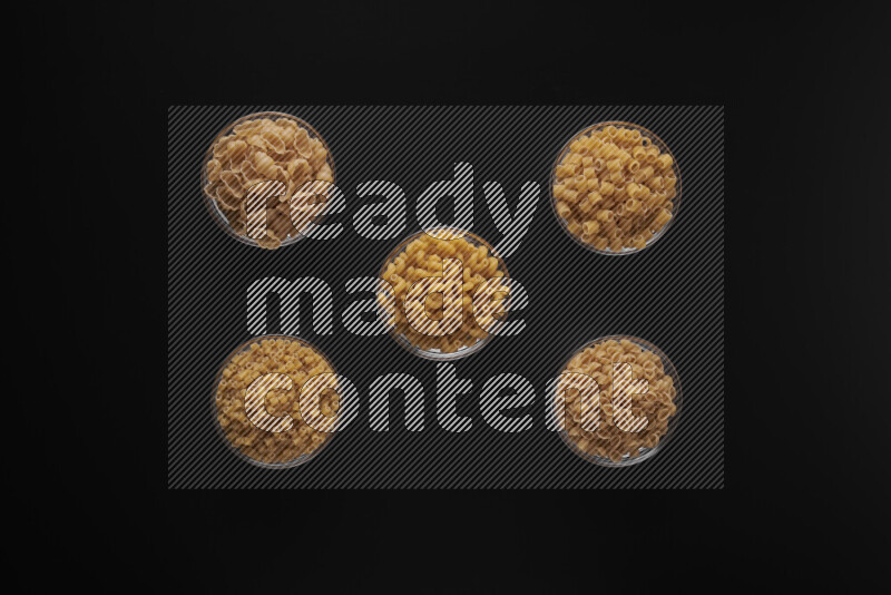 Different pasta types in 5 glass bowls on black background