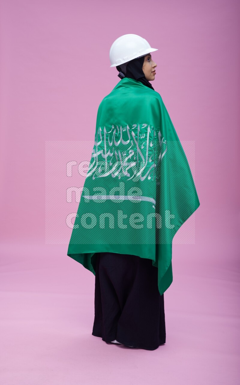 Saudi woman wearing Abaya with engineer vest and helmet standing holding Saudi flag on pink background