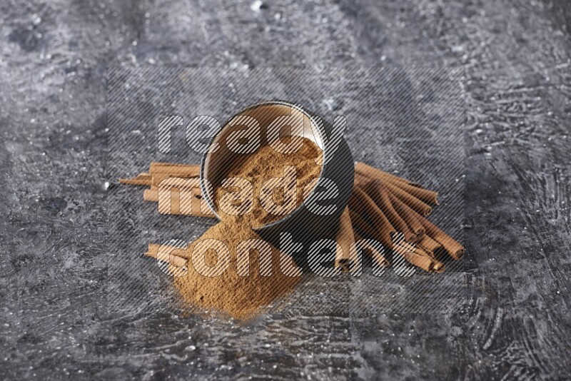 Black pottery bowl over filled with cinnamon powder and cinnamon sticks around the bowl on a textured black background