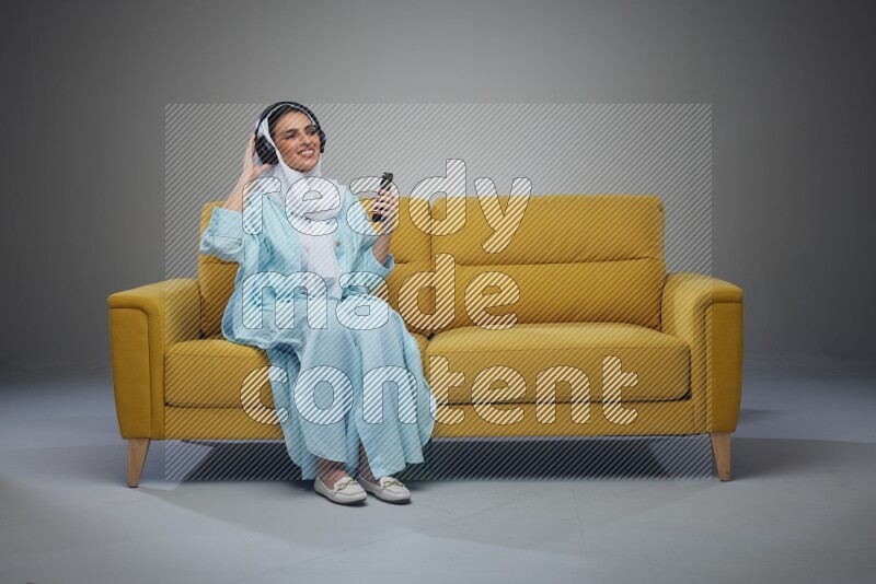 A Saudi woman wearing a light blue Abaya and a white head scarf sitting on a yellow sofa and holding an electronic while wearing headphone eye level on a grey background