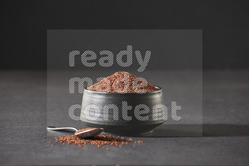 A black pottery bowl full of garden cress seeds and metal spoon full of the seeds on a black flooring