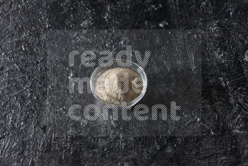 A glass bowl full of white pepper powder on textured black flooring