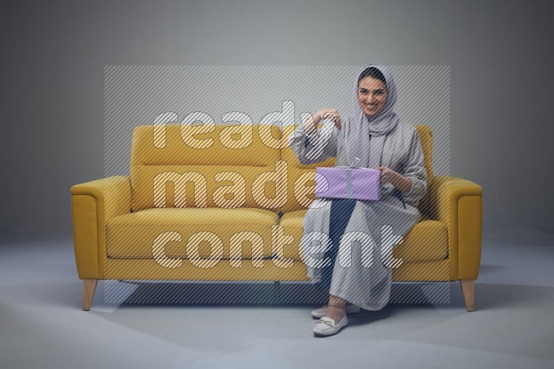 A Saudi woman wearing a light gray Abaya and head scarf sitting on a yellow sofa holding a gift box beside multi color gift boxes eye level on a grey background