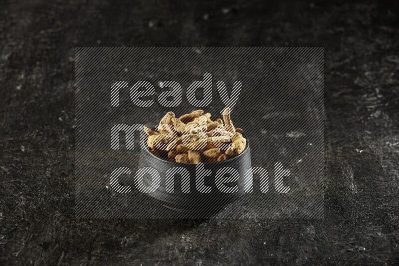 A black pottery bowl full of dried turmeric whole fingers on textured black flooring