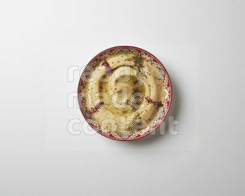 Hummus in a red plate with patterns garnished with zaatar & sumak on a white background