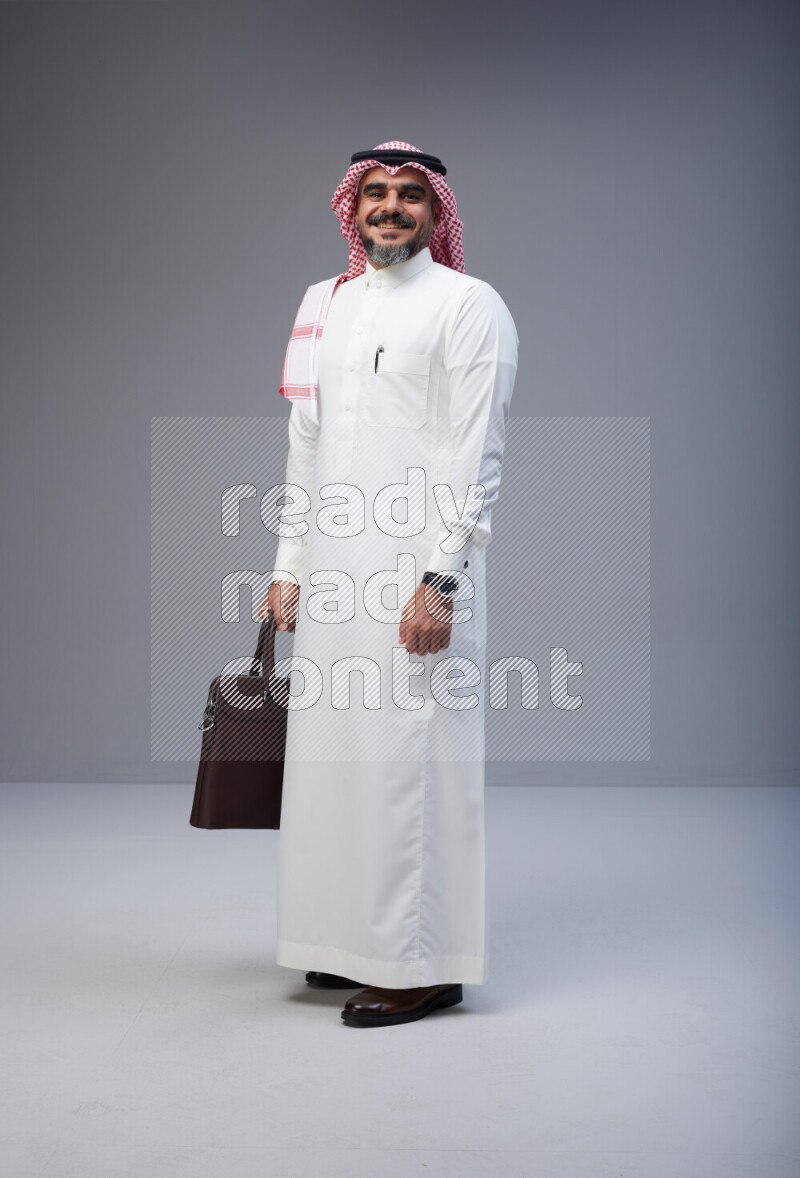 Saudi man Wearing Thob and red Shomag standing holding bag on Gray background