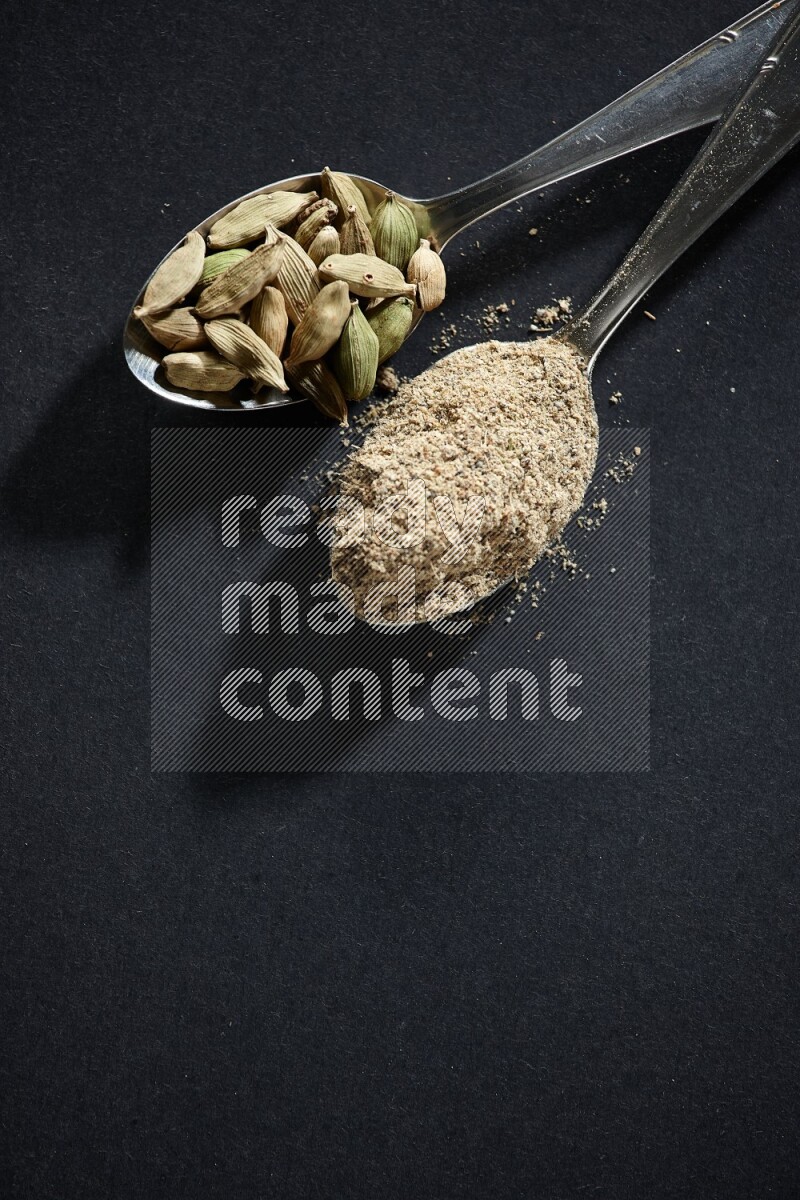 ملعقتان معدنيتان ممتلئتان ببودرة الهيل وحبوب الهيل على خلفية سوداء