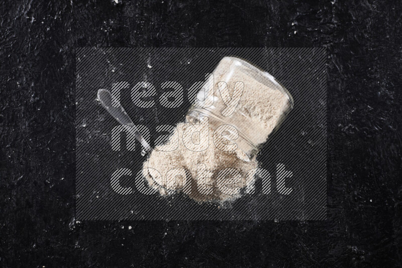 A glass jar full of onion powder flipped with some spilling powder on black background