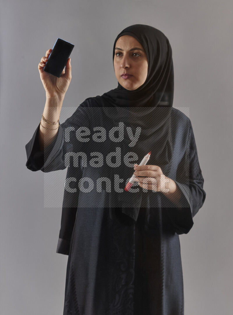 Woman in black abaya and hijab holding a marker pen to write on a transparent board with different reactions and expressions on grey background