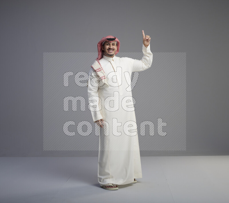 A Saudi man standing wearing thob and red shomag interacting with the camera on gray background