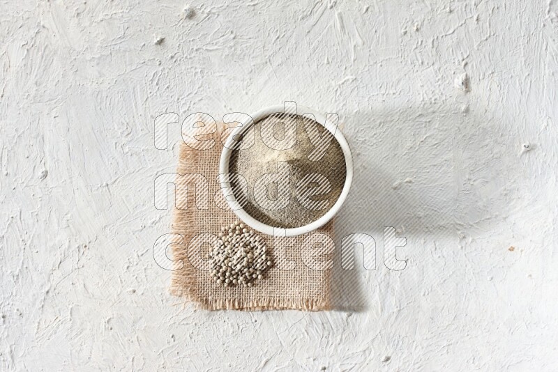 White pottery bowl full of white pepper powder set on a burlap piece of fabric with pepper beads and wooden pepper grinder on textured white flooring
