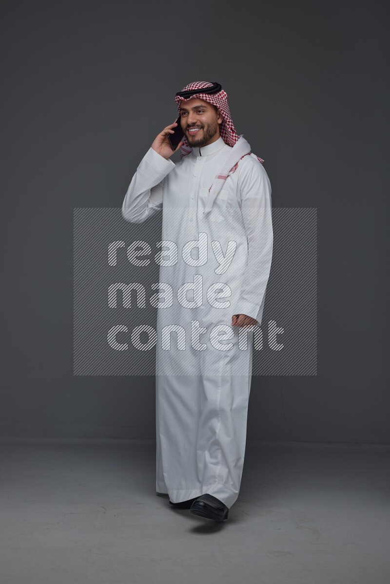 A Saudi man wearing Thobe and Shmagh talking in the phone eye level on a gray background