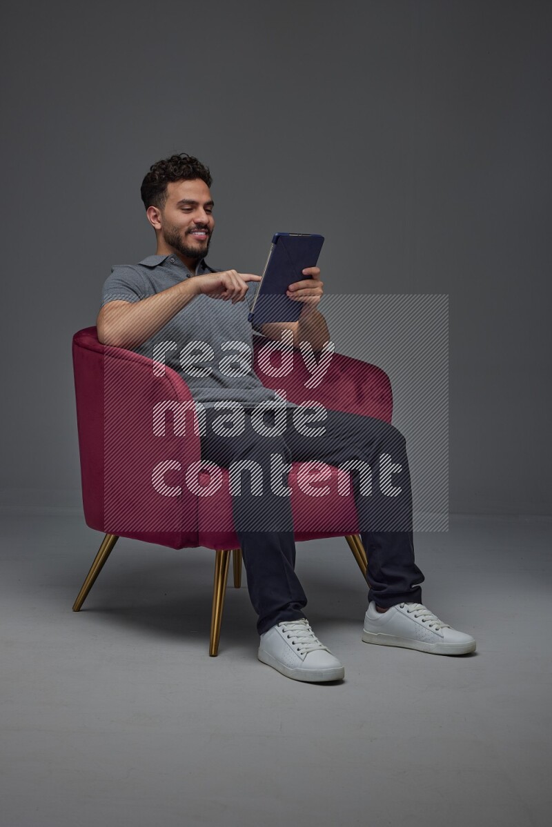 A man wearing casual and using his tablet while sitting on a burgundy chair eye level on a gray background