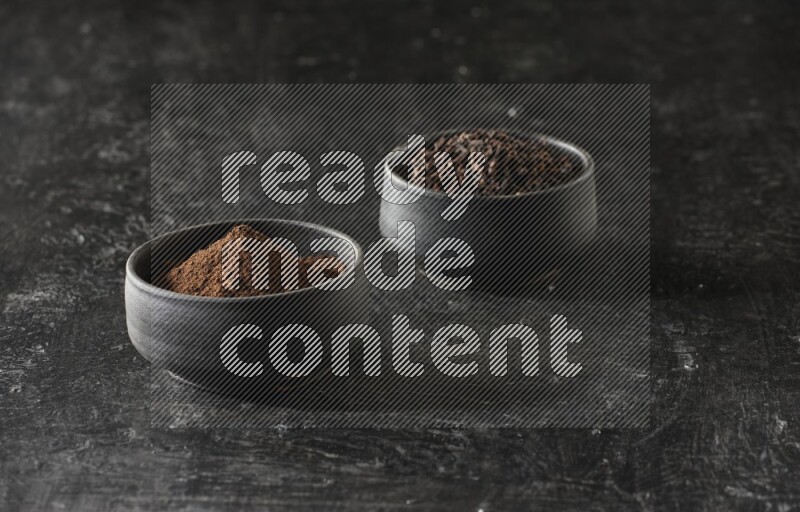 2 Black pottery bowls full of cloves and the other full of cloves powder on textured black flooring