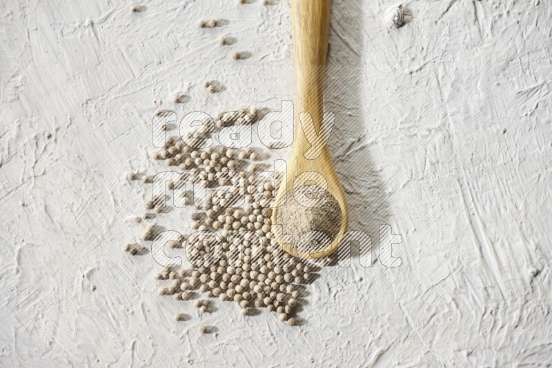 A wooden spoon full of white pepper powder and white pepper beads on textured white flooring