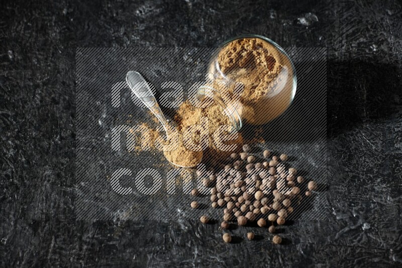 A flipped glass spice jar and metal spoon full of allspice powder and powder spilled out of it with whole balls on a textured black flooring