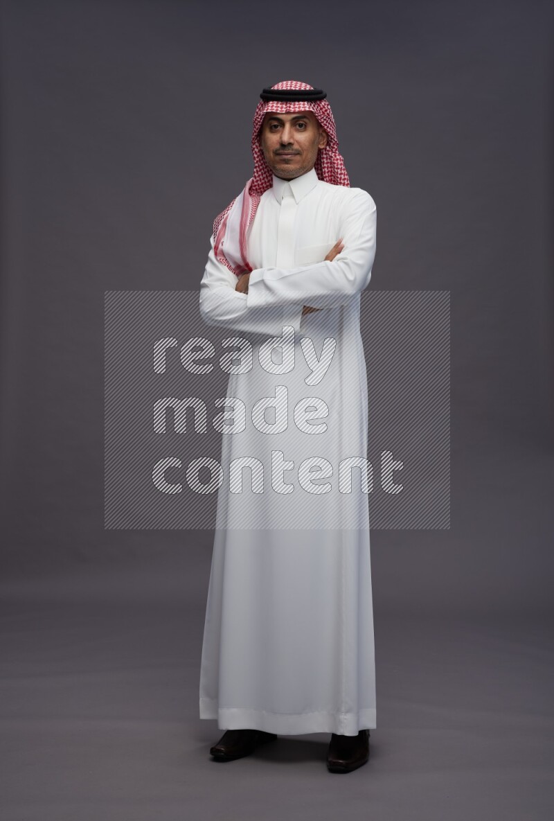 Saudi man wearing thob and shomag standing with crossed arms on gray background