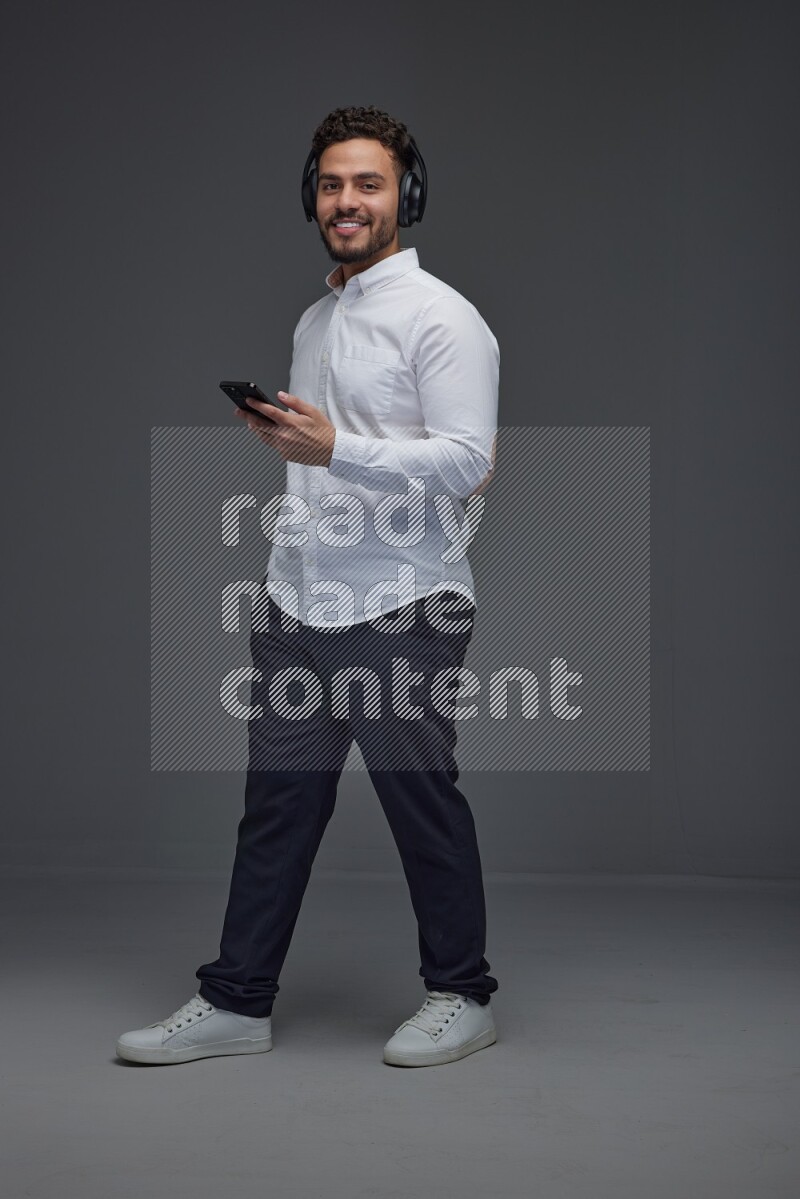 A man wearing smart casual and using his phone and headphone eye level on a gray background