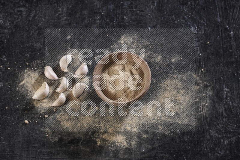 A wooden bowl full of garlic powder with some garlic cloves beside it on a textured black flooring