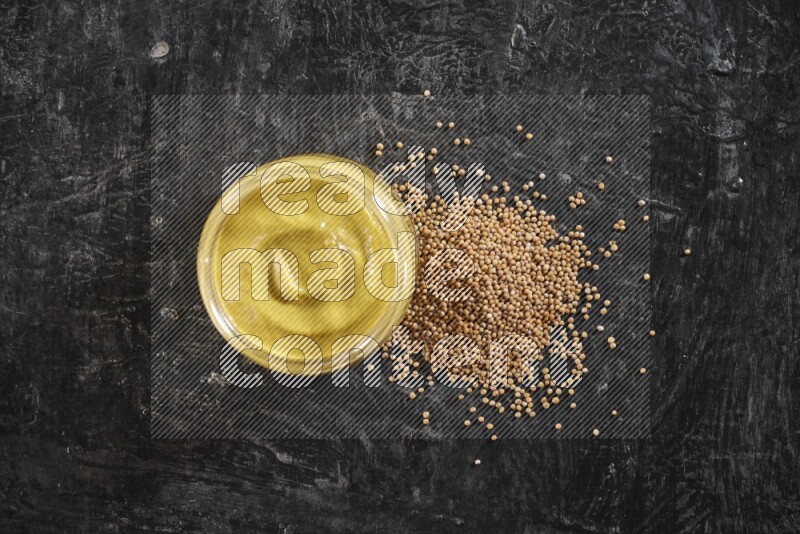 A glass jar full of mustard paste and mustard seeds spread next to it on a textured black flooring