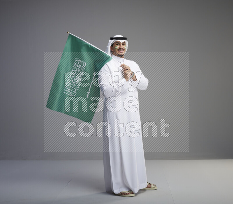 A Saudi man standing wearing thob and white shomag with face painting raising big Saudi flag on gray background