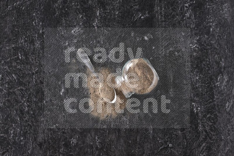 A flipped glass spice jar full of black pepper powder with a metal spoon full of powder on textured black flooring