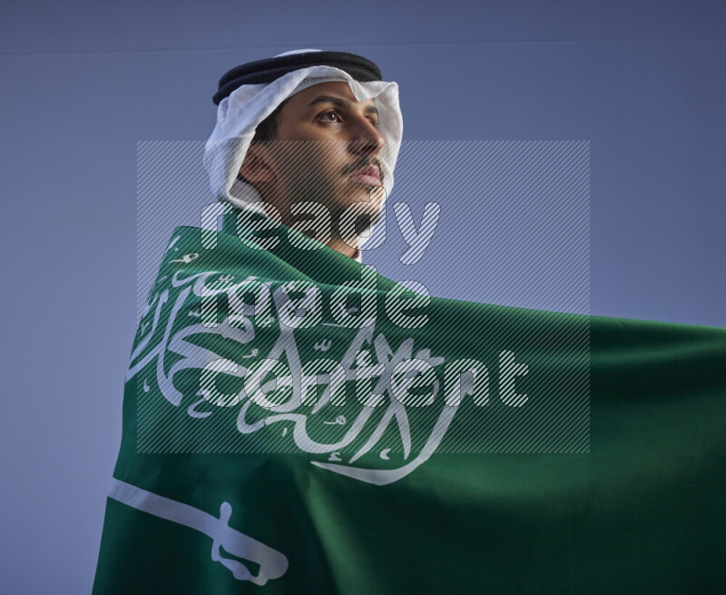 A close-up shot of Saudi man wearing thob and white shomag wrapping big Saudi flag on gray background